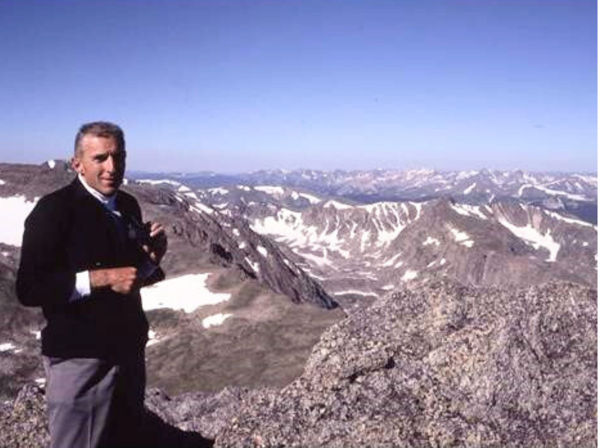 Dale Hein stands on the top of a mountain with a mountain range behind him.