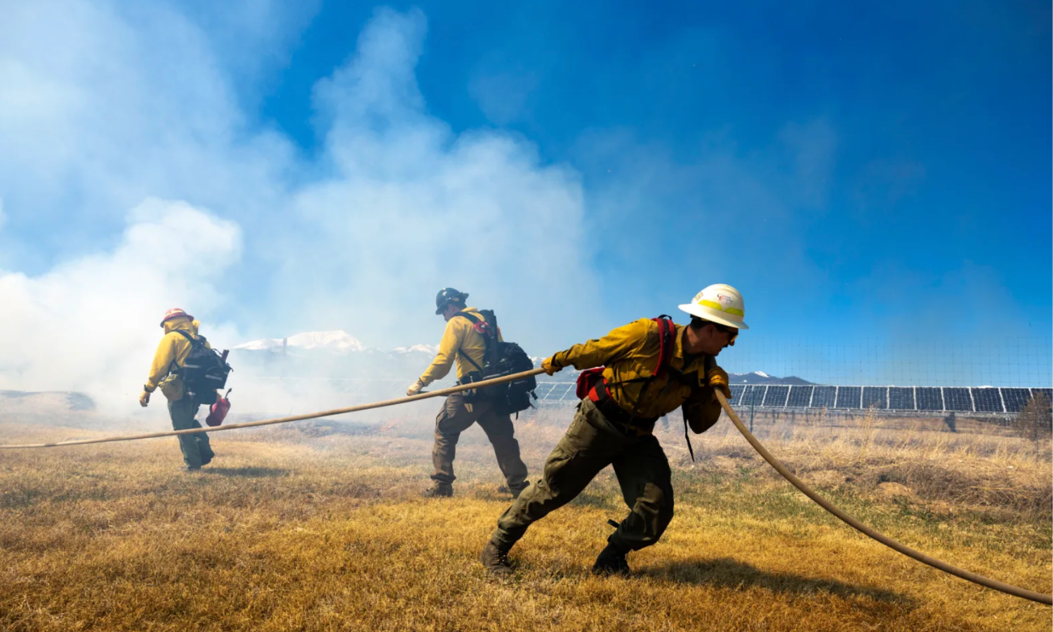 firefighters pull a hose while smoke billows in the background