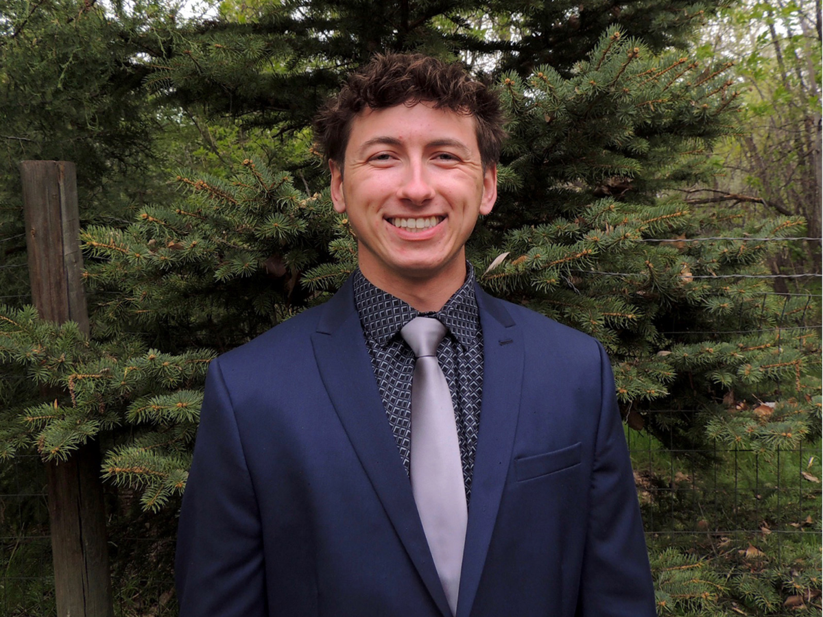 Noah Niemeyer smiles while wearing a blue suit jacket in front of a pine tree.