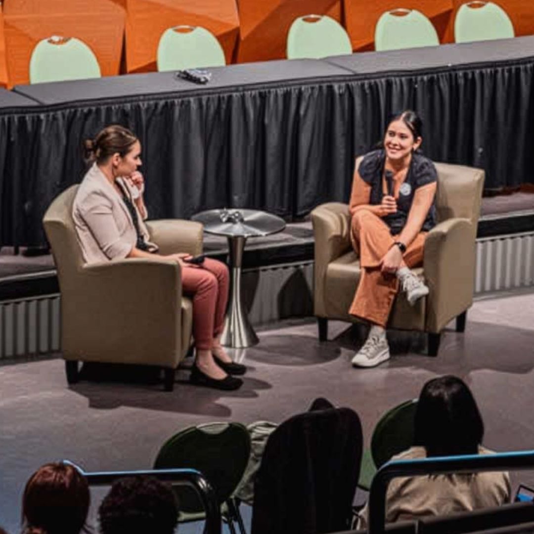 Two people sit in chairs on a stage talking at The Symposium