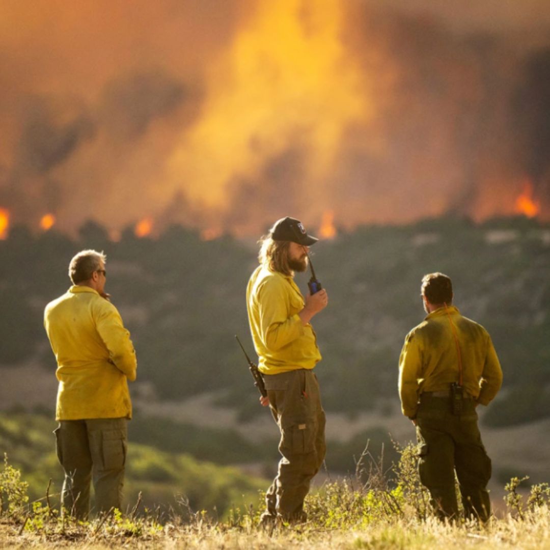 Three firefighters stand in front of a blazing wildfire. 