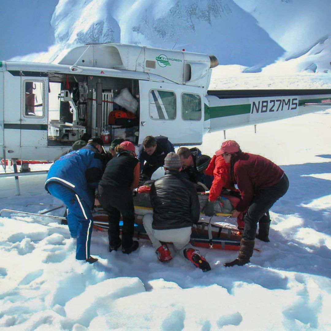 A group of people put someone on a stretcher on top of a snowy mountain. 
