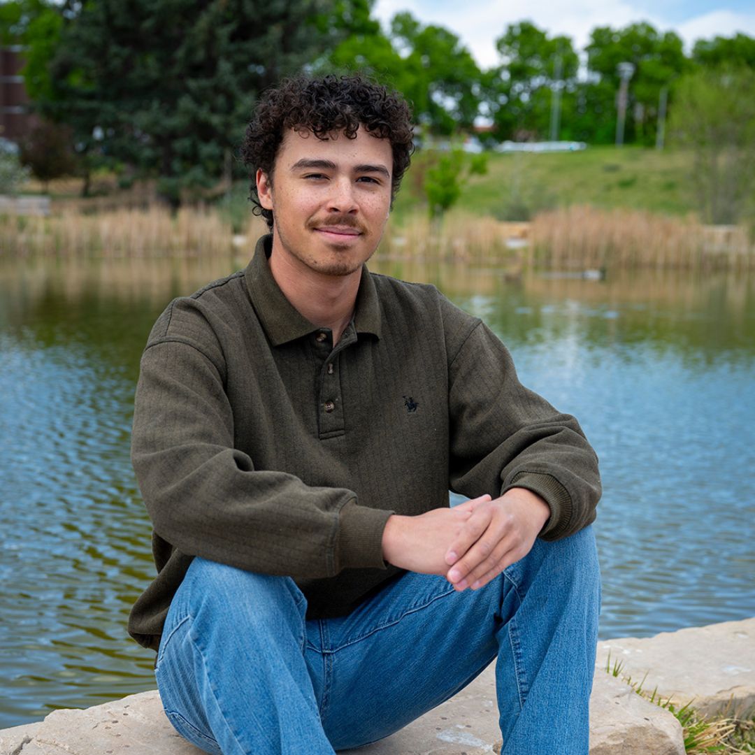 A photo of Alex Wagner sitting on rocks with a pond in the background.
