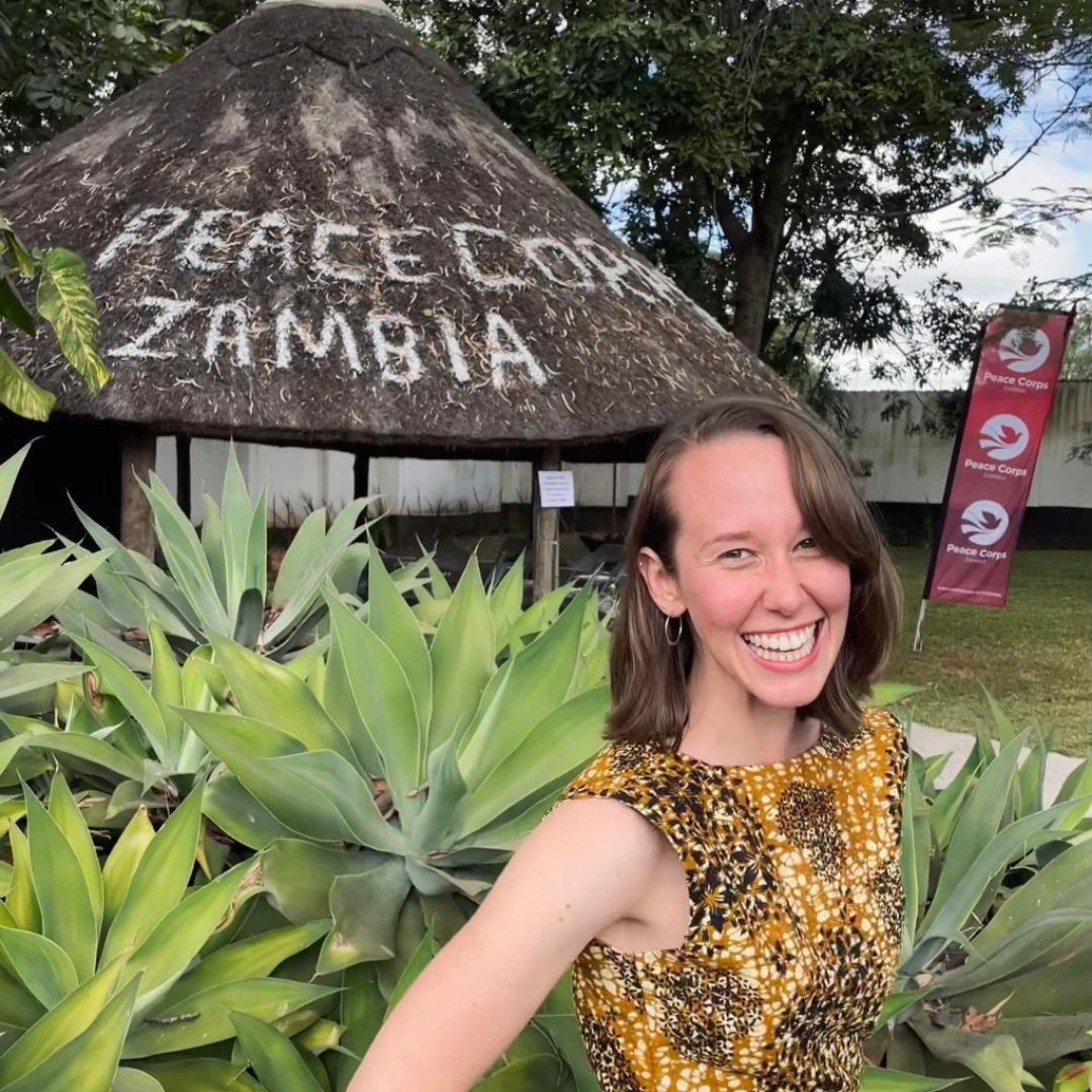 A student stands in front of a hut that says 