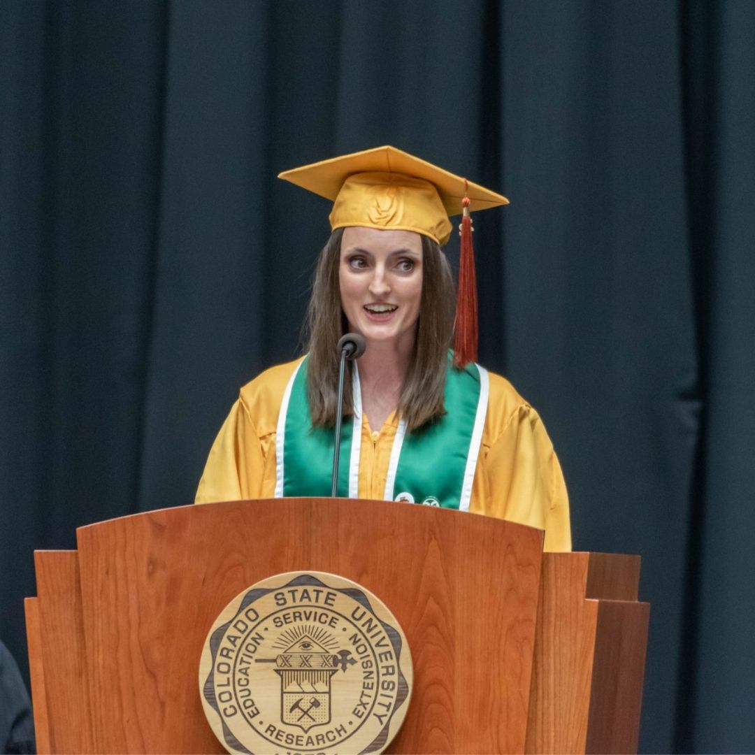 A student in a gold gown and mortar board speaks into a microphone at commencement. 
